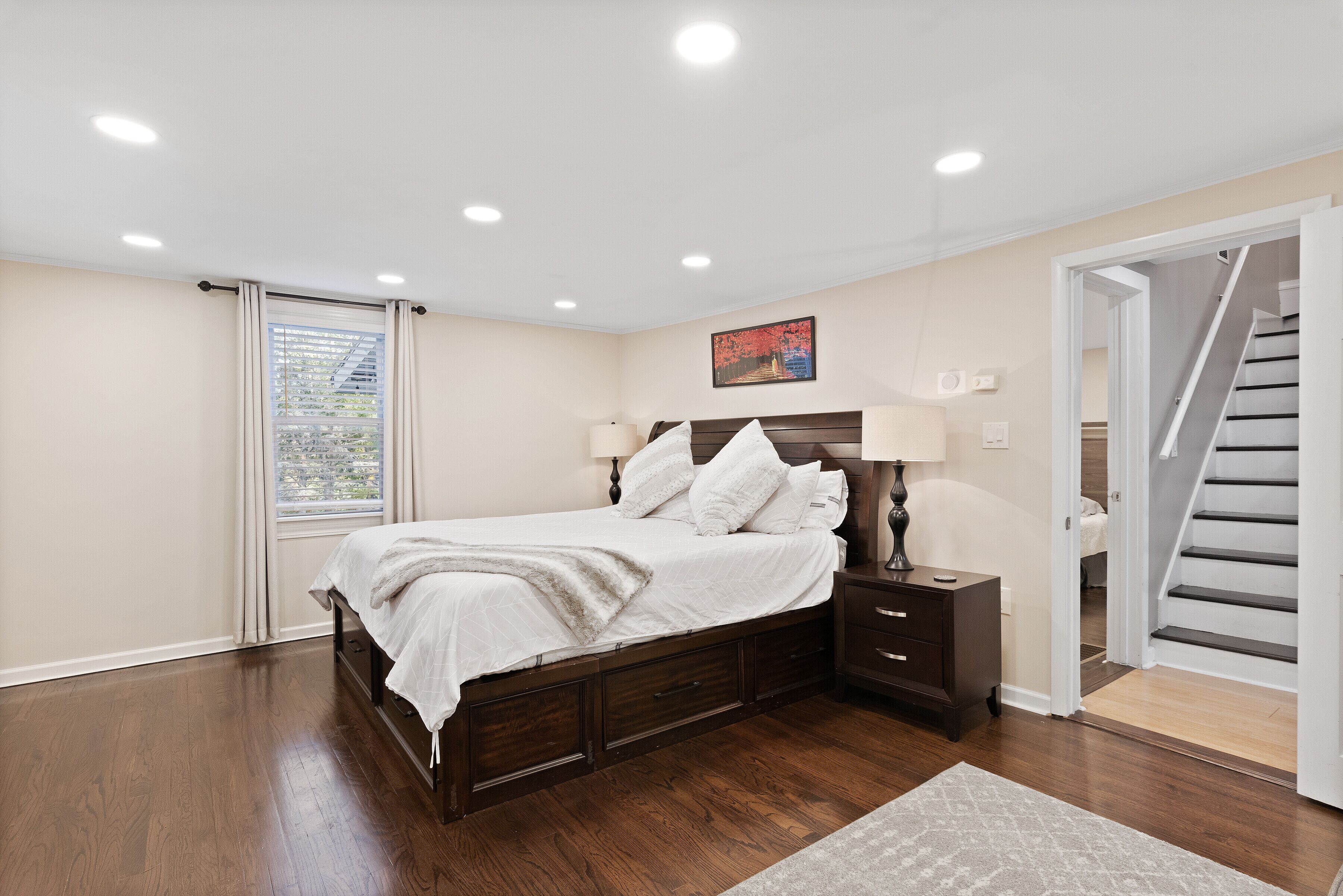 A bedroom with a large bed, wooden headboard, two bedside tables, a window with curtains, and a staircase to the right.