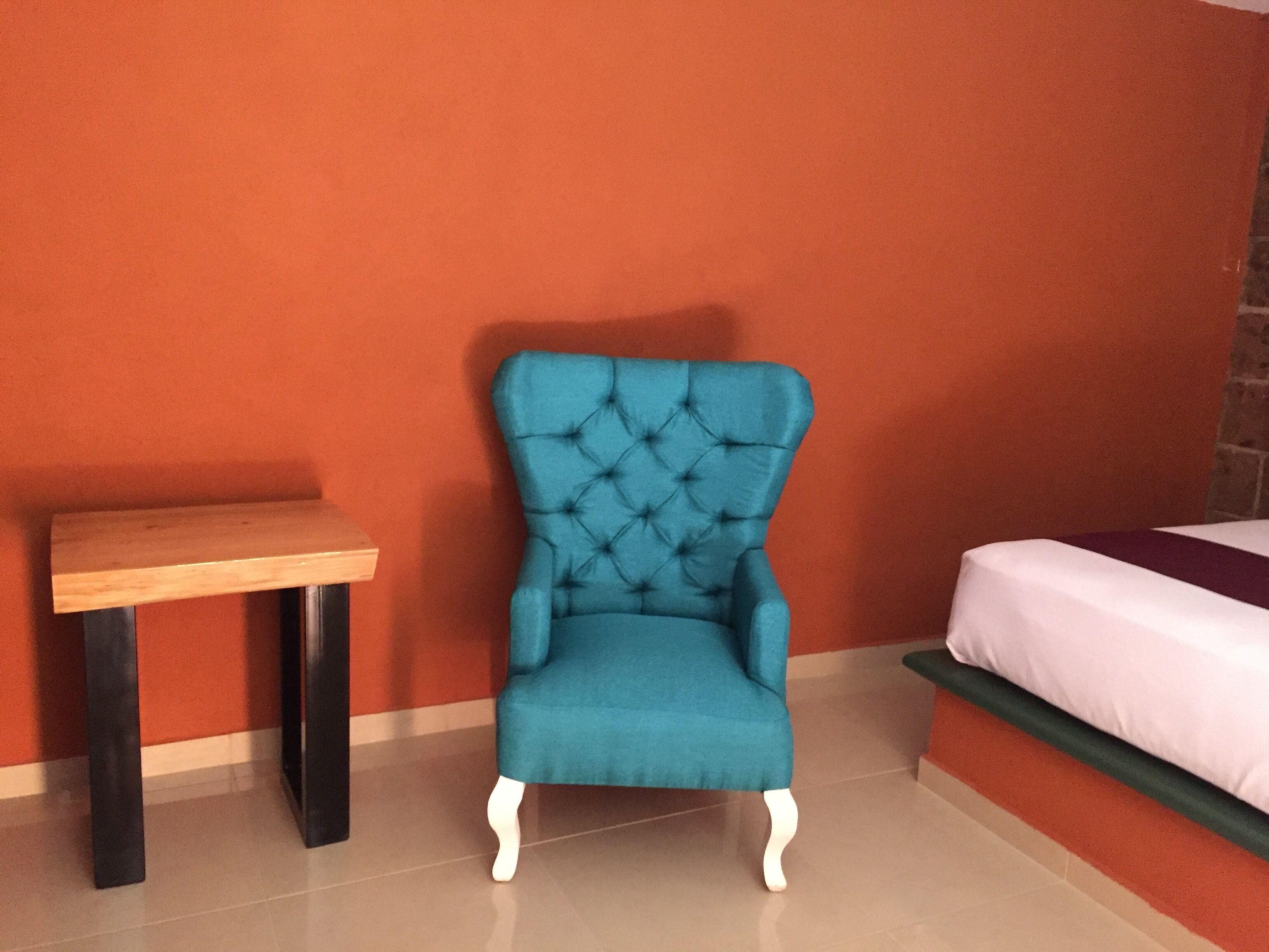 A teal armchair with a tufted backrest, a wooden side table, and a bed with a white and green bedspread in a room with orange walls and a tiled floor.