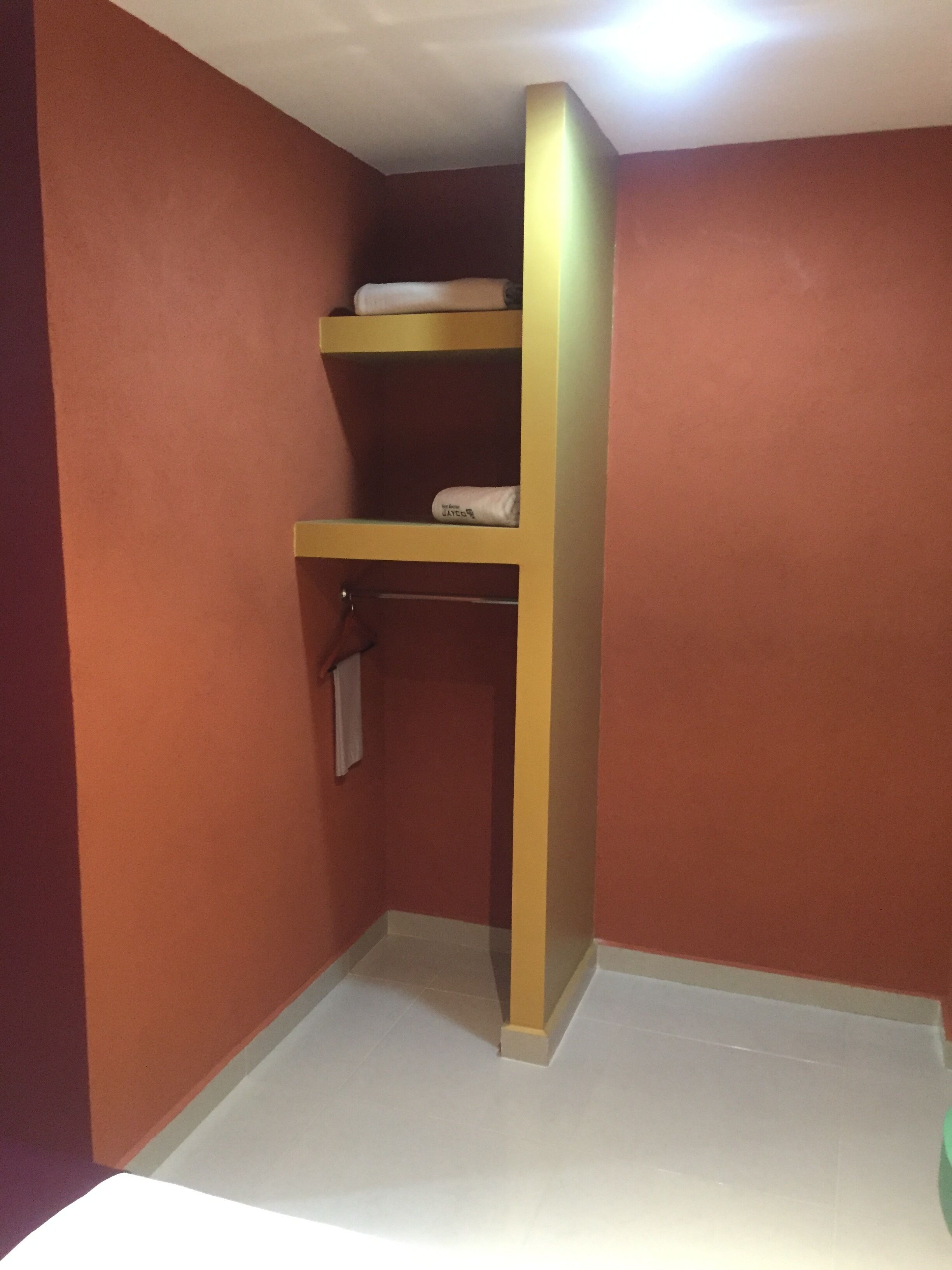 A small, enclosed room with a single wooden shelf unit against the wall, a white towel hanging on a metal rod, and a white floor.