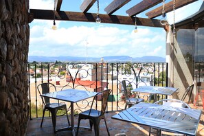 Une terrasse avec des tables et des chaises en métal, un mur de pierre et une vue sur des bâtiments et des montagnes situés au loin.