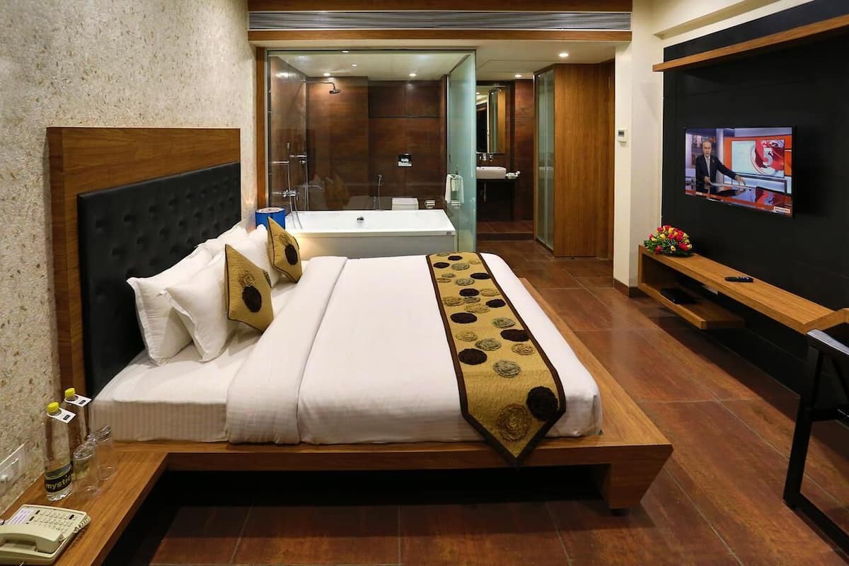 Quarto de hotel moderno com uma cama grande, uma televisão, banheiro e um banco de madeira.