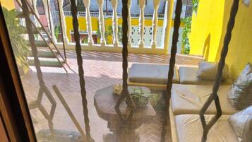 Una habitación con ventana que da a un patio con pared amarilla, baranda blanca y una zona de estar con cojines.