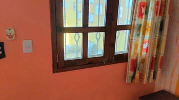 Ein Zimmer mit Fenster, Tür, Fliesenboden, einem kleinen Holzschrank und einem Bett mit gemustertem Überwurf.