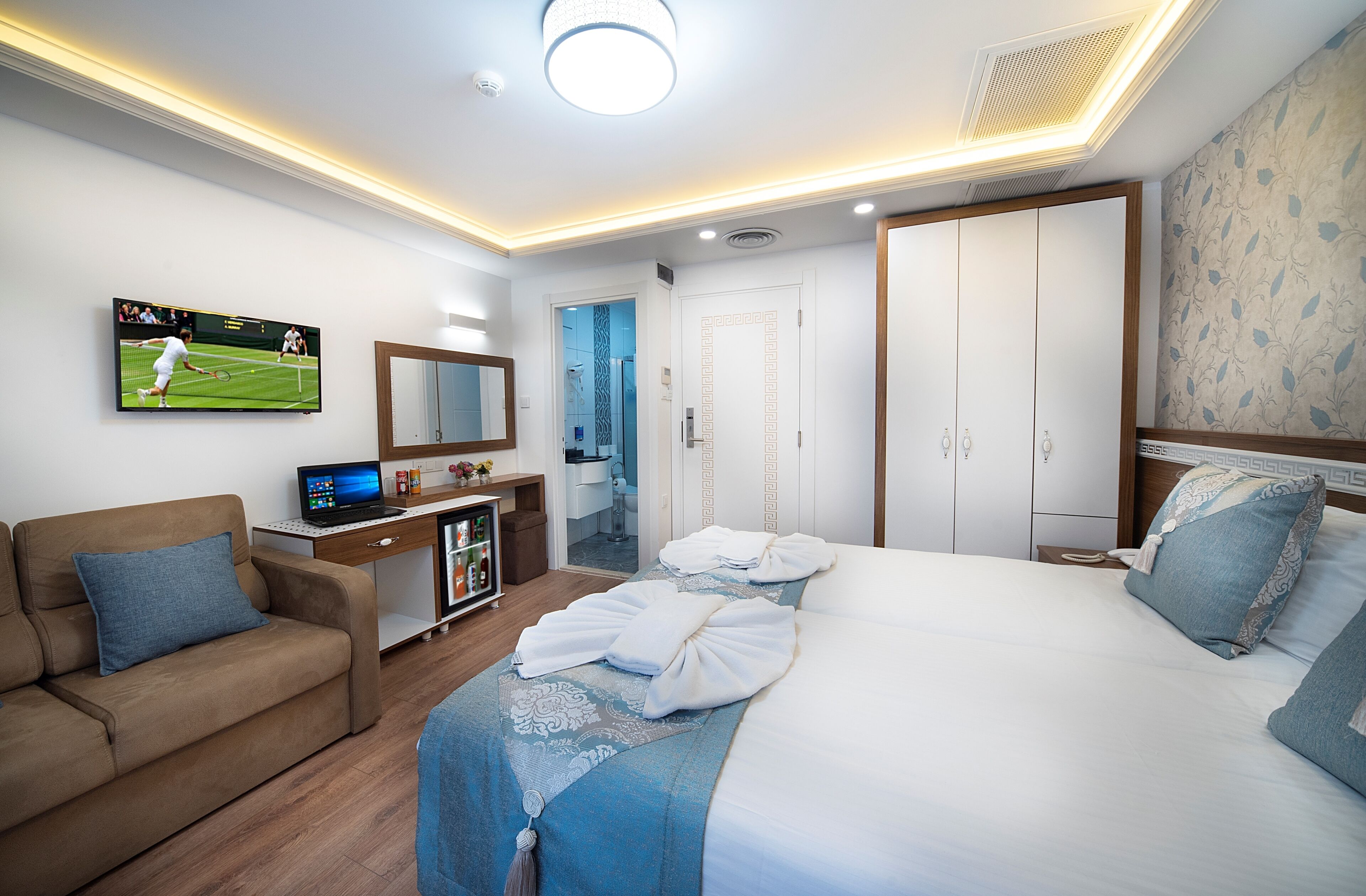 Een hotelkamer met een bed, een slaapbank, een bureau met een laptop, een televisie waarop een tenniswedstrijd wordt vertoond, en een kledingkast.