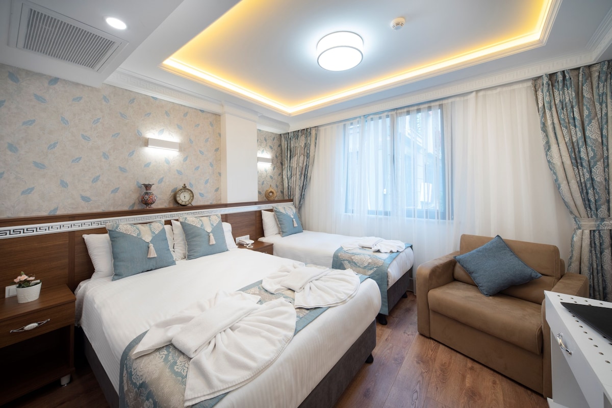 A hotel room with two beds, a sofa, a desk, and a large window with curtains.