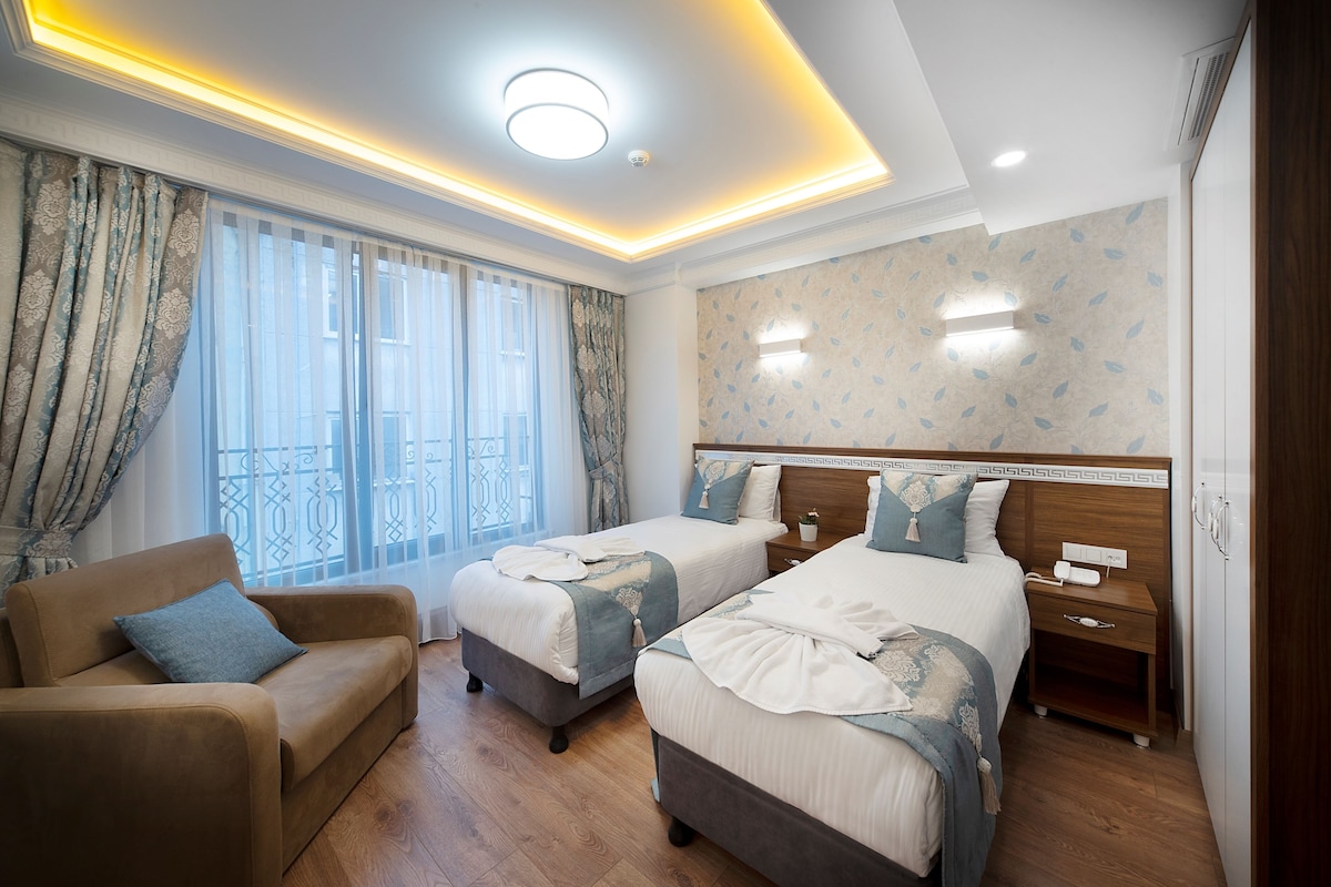 A hotel room with two beds, a sofa, a nightstand, and a window with curtains.