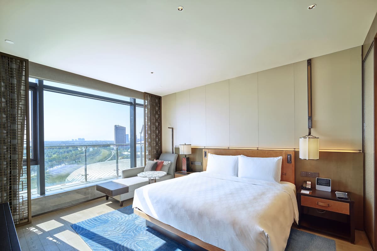 Habitación de hotel moderna con una cama grande, zona de estar y vistas a un paisaje urbano.