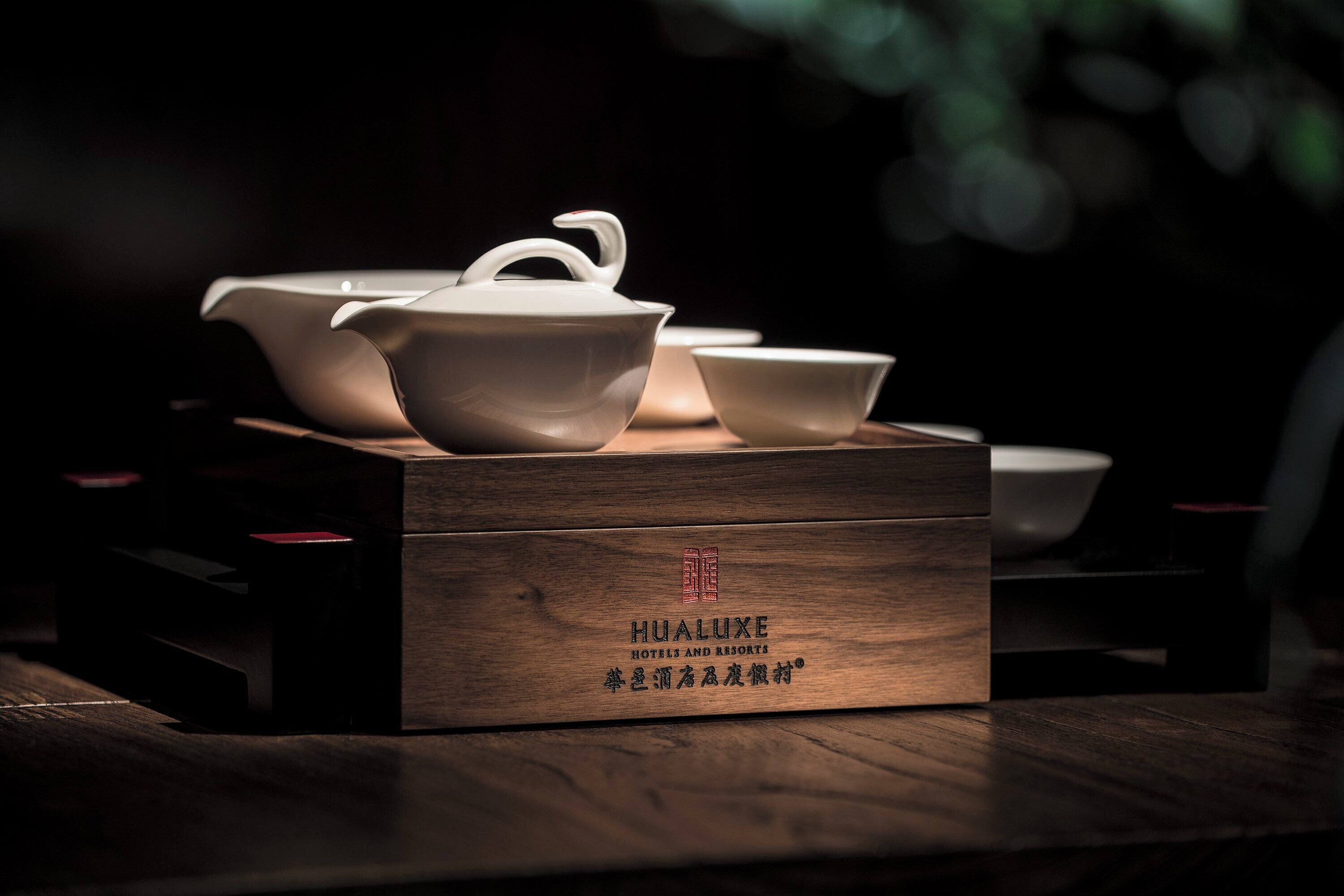 Una caja de madera con la inscripción 'HUALUXE HOTELS AND RESORTS' y caracteres chinos, que contiene una tetera, una taza y un platito sobre una mesa.