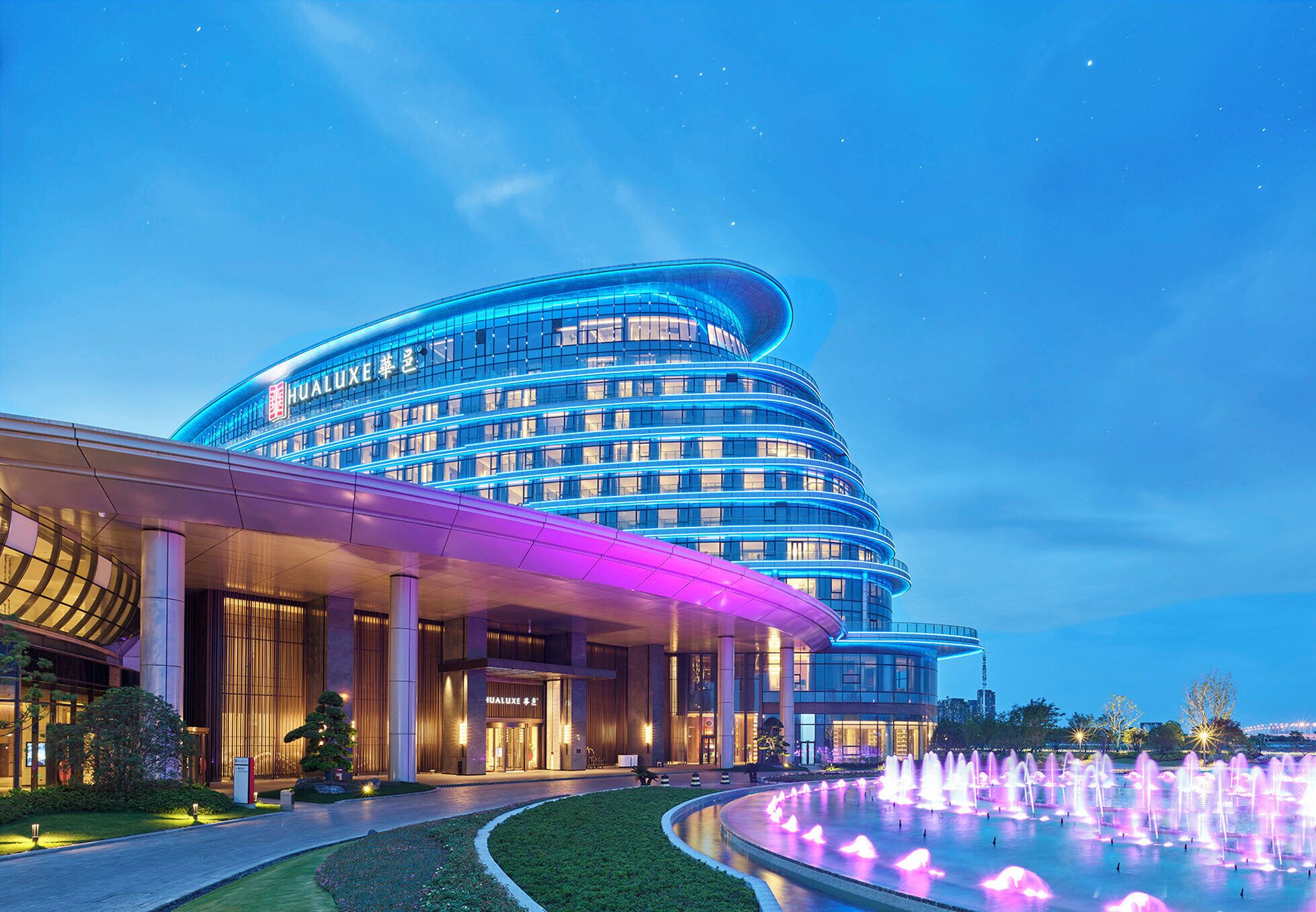 A modern hotel with a curved facade, illuminated by blue lights, and a fountain in the foreground.