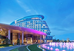 Un hôtel moderne à la façade incurvée, illuminée de lumières bleues, avec une fontaine en premier plan.