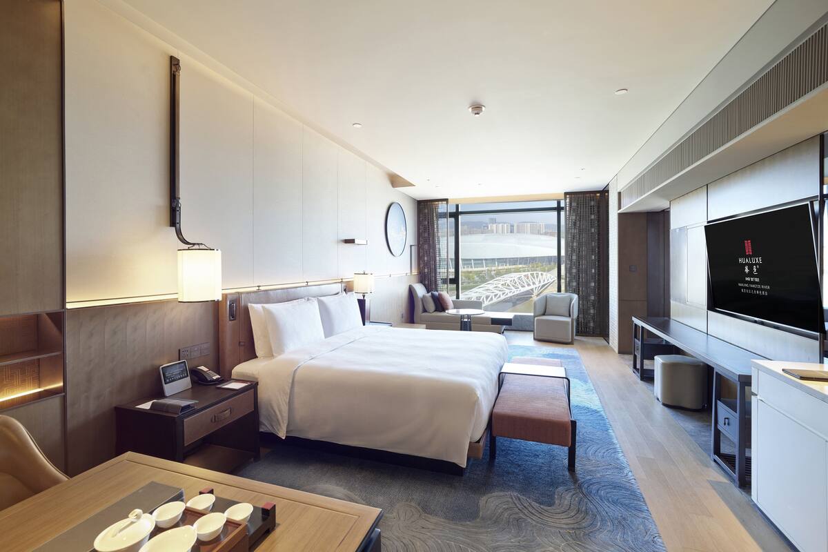 Una habitación de hotel moderna con una cama grande, mesitas de noche, un escritorio con computadora, televisor y vistas a un puente.