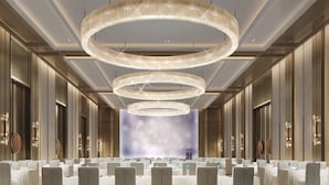 Een congreszaal met meerdere tafels en stoelen, grote ronde kroonluchters en een podium met achtergrond.