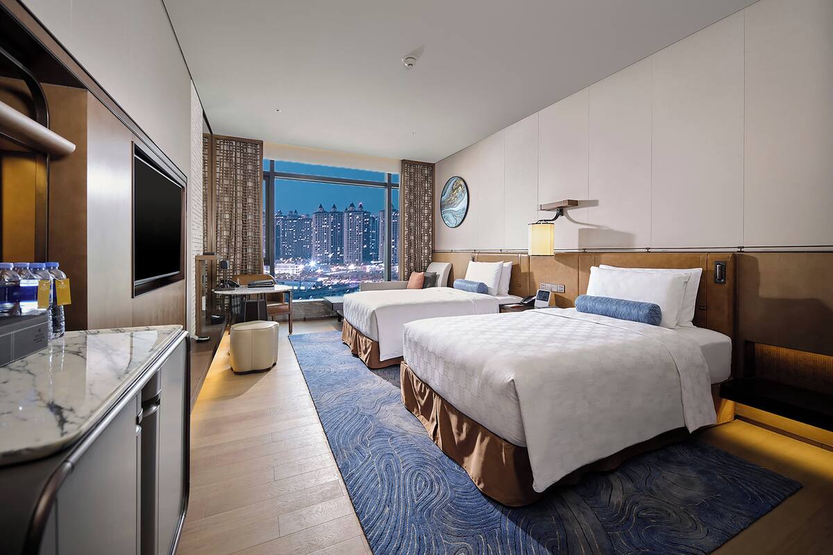 Habitación de hotel con dos camas, un escritorio, televisión y vistas a la ciudad.