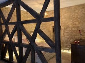 A rustic room with exposed wooden beams, a stone wall, a bed with a white bedspread, and a small table with candles.