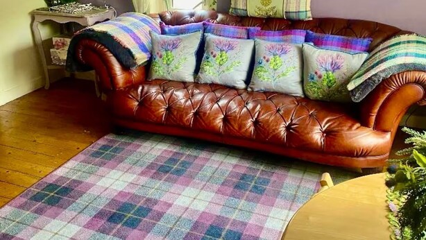 Un sofà de cuir marró amb coixins florals, una catifes escoceses i una paret amb una decoració metàl·lica circular.