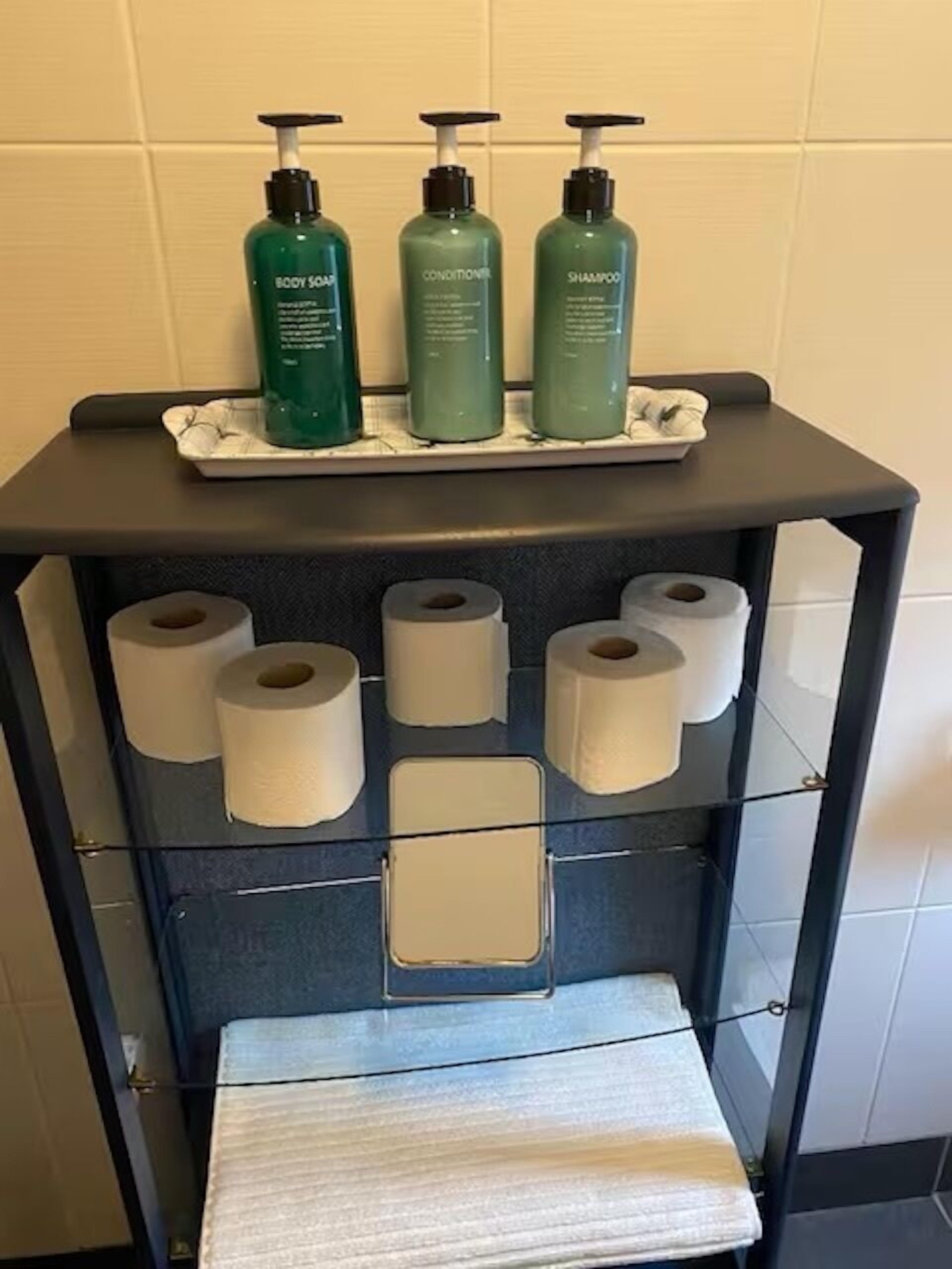Een badkamer met een plank waarop drie flessen groene vloeibare zeep staan en een rek met meerdere rollen toiletpapier.