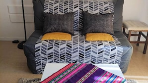 A sofa with a geometric patterned cover, two mustard-colored cushions, and a colorful woven throw on a white table.