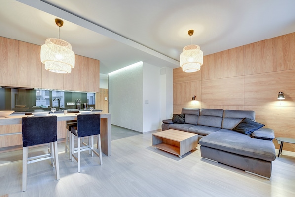A modern living room with a grey sectional sofa, a coffee table, and a kitchen area with a dining table and bar stools.