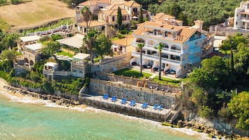 An aerial view of a coastal property with multiple buildings, a stone wall, and a beach with blue lounge chairs.