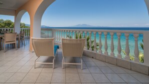 Un balcone con due poltrone in vimini, un tavolo e vista sul mare e su una montagna.