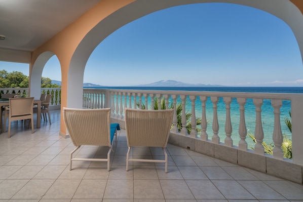 A balcony with two wicker chairs, a table, and a view of the sea and a mountain. - Playa Del Zante - 2 Bedroom Sea View Apartment, Playa del Zante (Zakynthos)