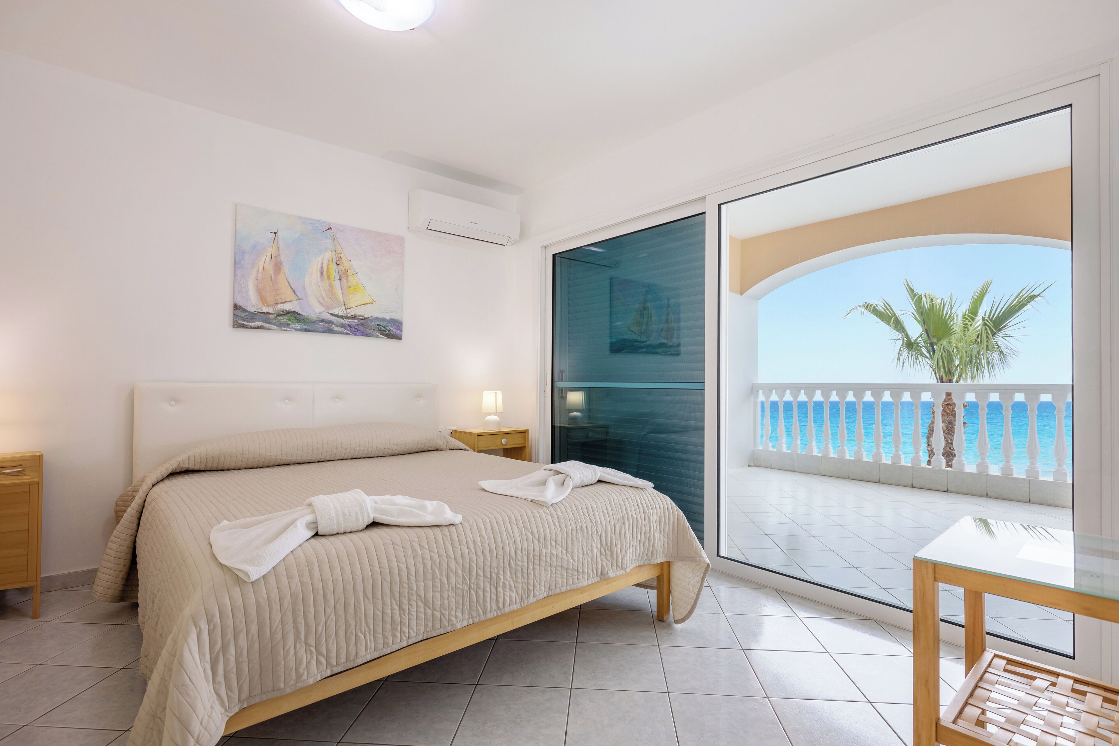A bedroom with a bed, a nightstand, a bedside table, a glass door leading to a balcony with a view of the sea and palm trees.