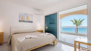 A bedroom with a bed, a nightstand, a bedside table, a glass door leading to a balcony with a view of the sea and palm trees.