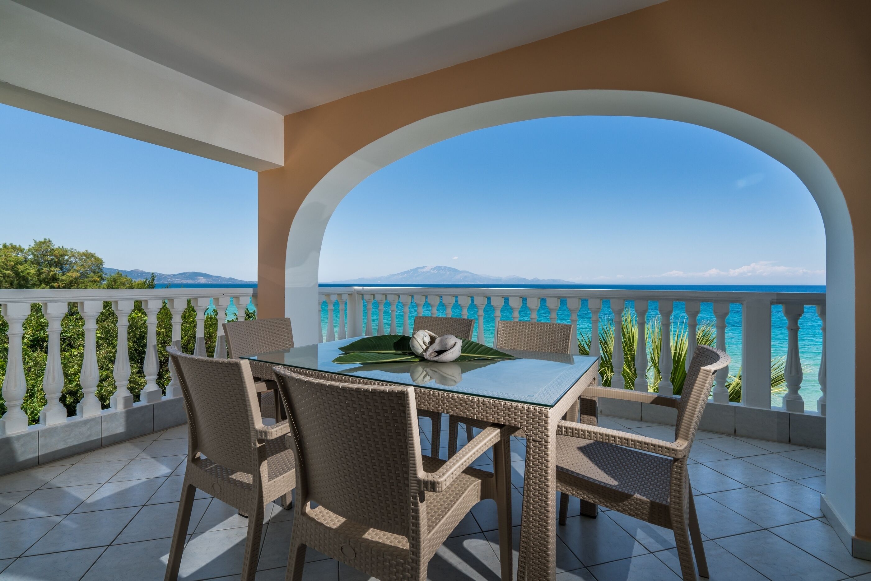 A balcony with a glass table and wicker chairs, overlooking a coastal view with mountains in the distance.