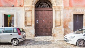 Ein Gebäude mit einer großen, verzierten Eingangstür, ein kleiner Laden mit einem grünen Kreuzschild und zwei parkende Autos.