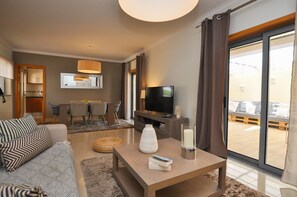 Ein modernes Hotelzimmer mit einer Sitzecke, einem Couchtisch mit Büchern, einem Fernseher und einem Balkon mit Blick.