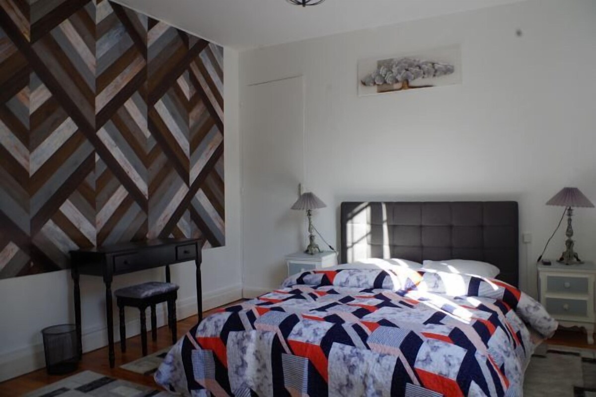 Une chambre à coucher comprenant un lit, deux tables de chevet, une chaise et un mur orné d’un motif géométrique.