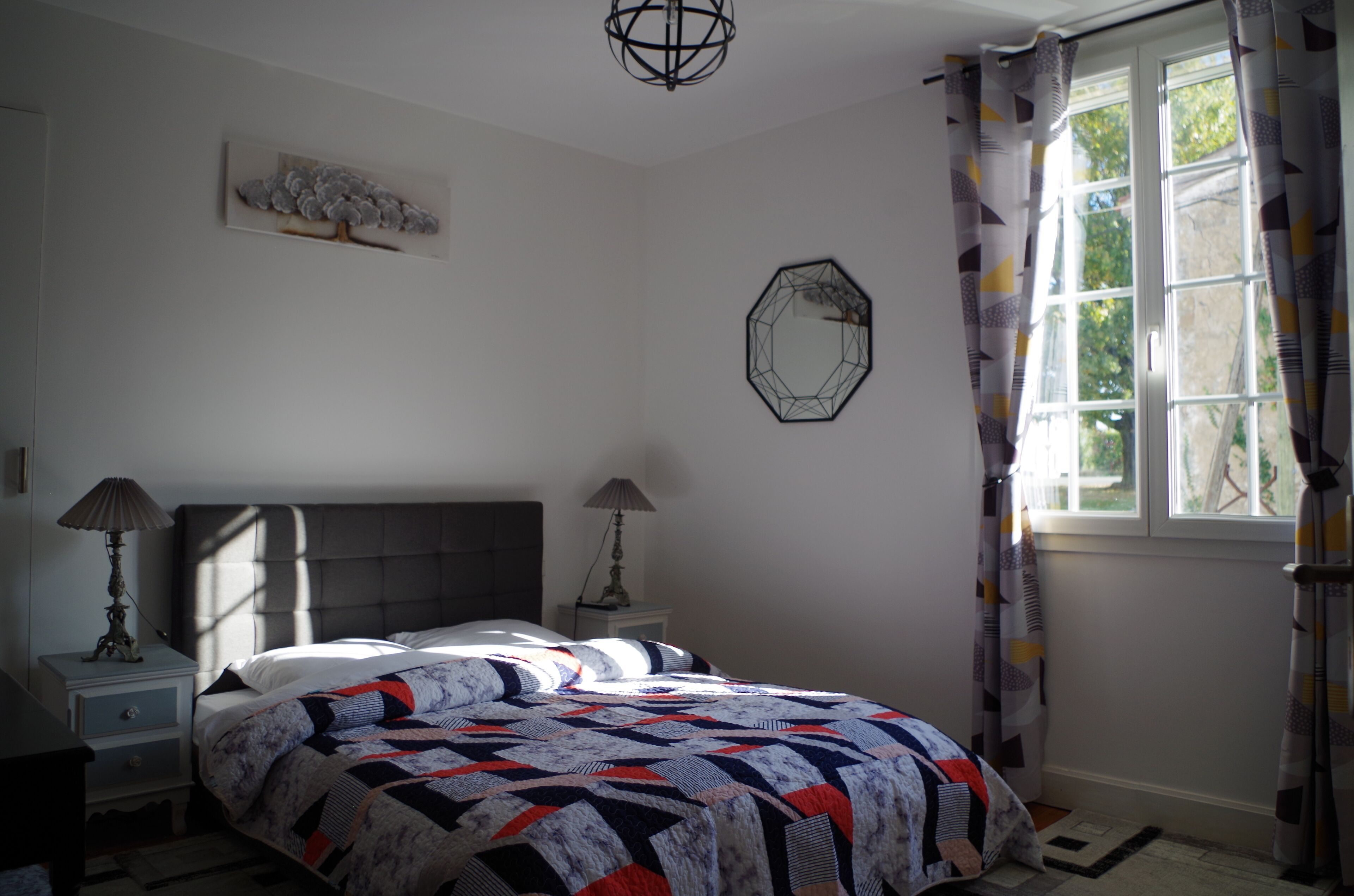 A bedroom with a bed, two bedside tables, a lamp, a wall clock, a window with curtains, and a geometric wall decoration.