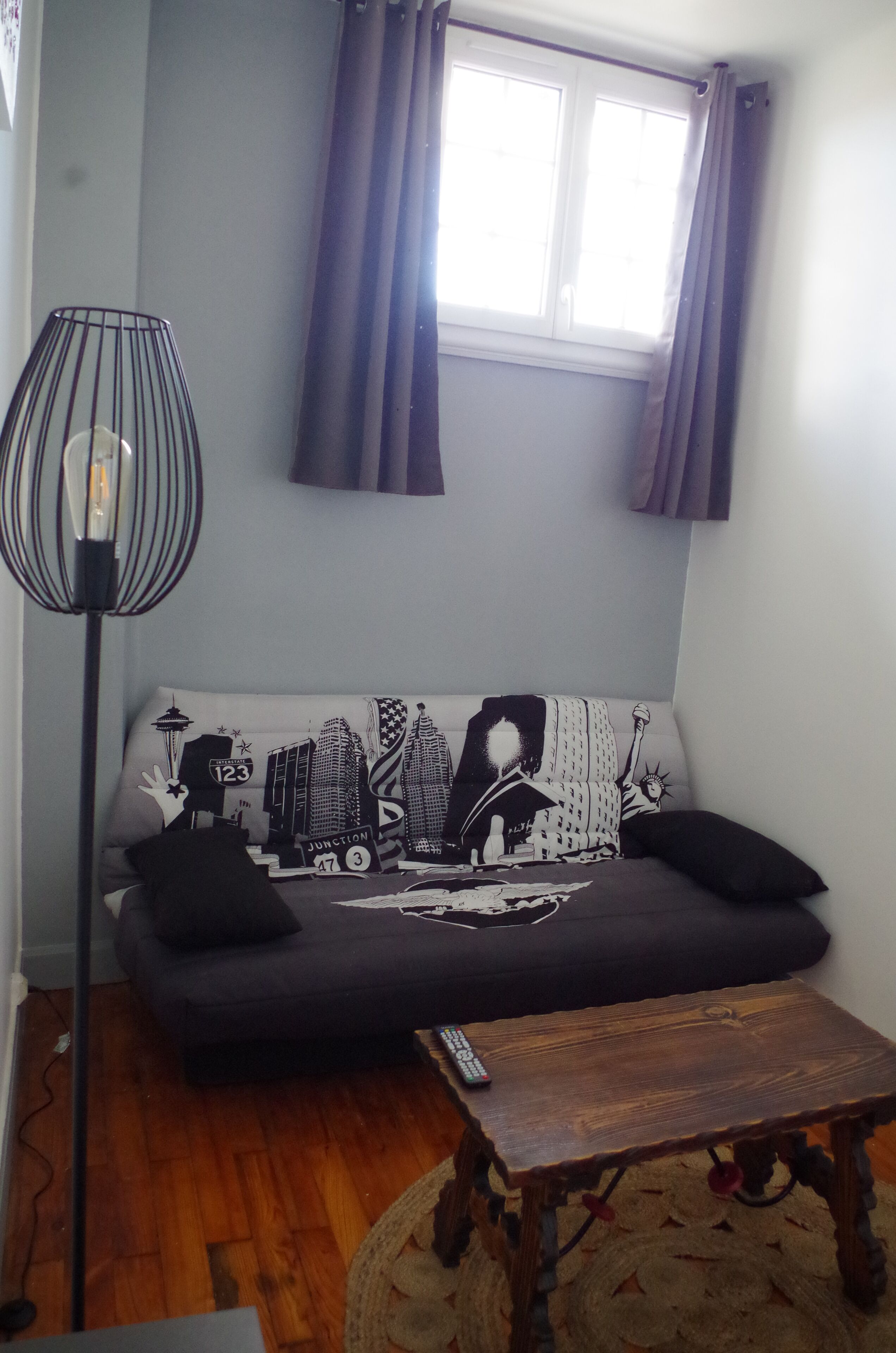 Una sala de estar con un sofá, una pequeña mesa de madera, una lámpara de pie y un ventanal con cortinas.