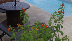 An outdoor pool area with a stone wall, a wooden table with chairs, and a large umbrella. - Maison Elizondoa (Masparraute)