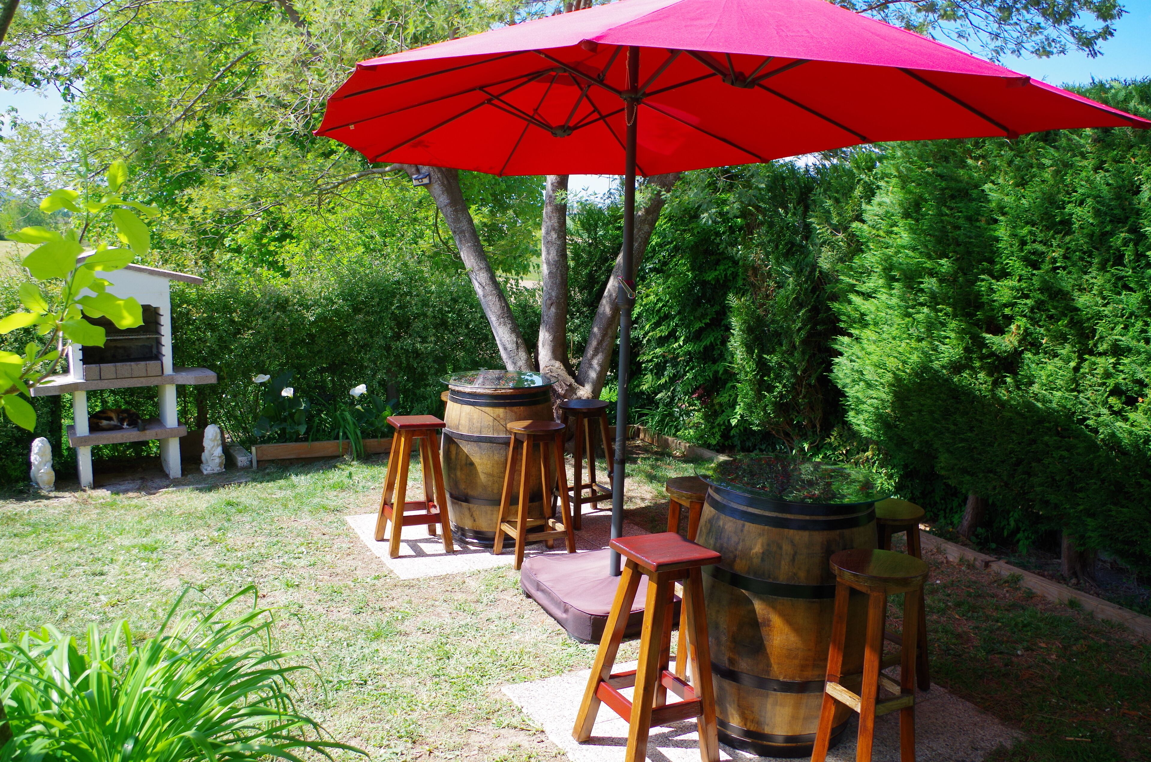 A patio area with wooden stools and barrels, surrounded by lush greenery.