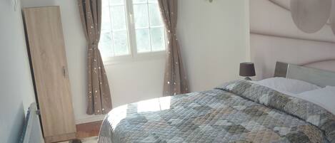 Un dormitorio con cama, radiador, ventana con cortinas, espejo en la pared y lámpara en el techo.