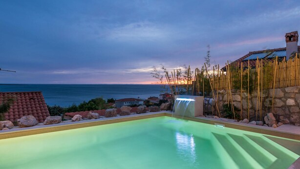 A swimming pool with a waterfall feature, surrounded by rocks and a bamboo fence, overlooking a coastal area with houses and a sunset.