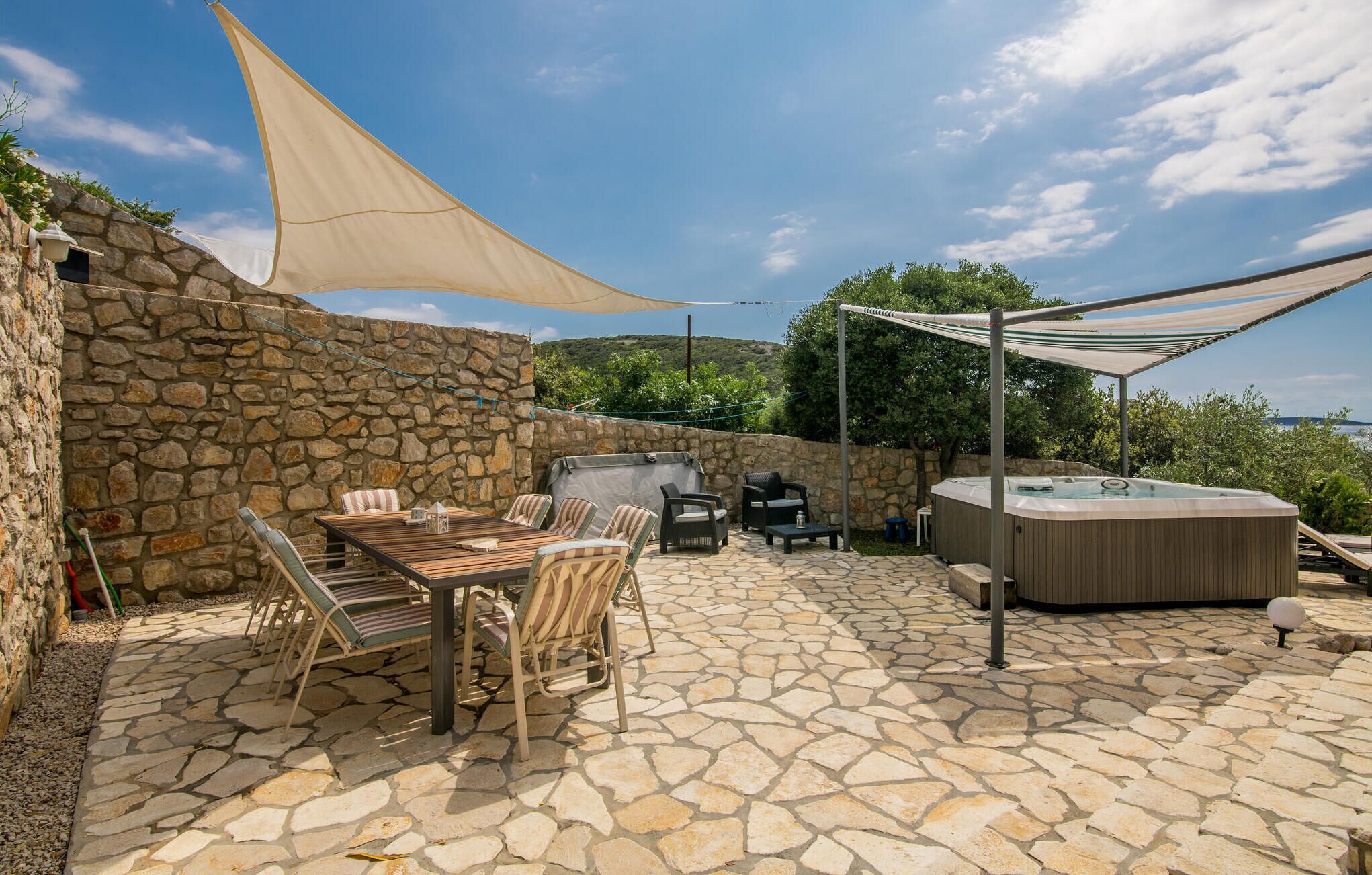 An outdoor patio with stone flooring, a wooden table with chairs, a hot tub, and a stone wall.