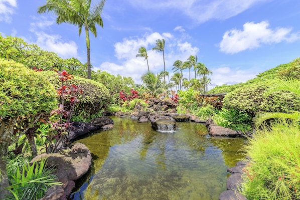 A serene garden with a small pond, lush greenery, and palm trees.