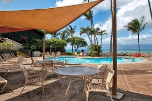 Un espace aménagé au bord de la piscine, avec des chaises longues et une table, entouré de palmiers et offrant une vue sur l’océan.