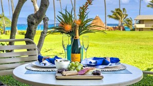 Ein Tisch gedeckt mit einer Champagnerflasche, Gläsern, einer Platte mit Trauben, Käse und einer Obstplatte, draußen mit Blick auf das Meer und Palmen.