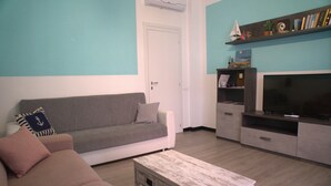 Un salon avec un canapé gris, une table basse en bois, un téléviseur à écran plat et une bibliothèque agrémentée d’objets décoratifs.
