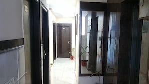 A hallway with a tiled floor, a door with a small potted plant, and a glass door reflecting the interior. - Hotel Green View Palace (Noida)