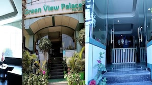 A hotel lobby with a sofa and a coffee table, a reception area with a staircase and potted plants, and a view of the entrance with a sign reading "Green View Palace". - Hotel Green View Palace (Noida)