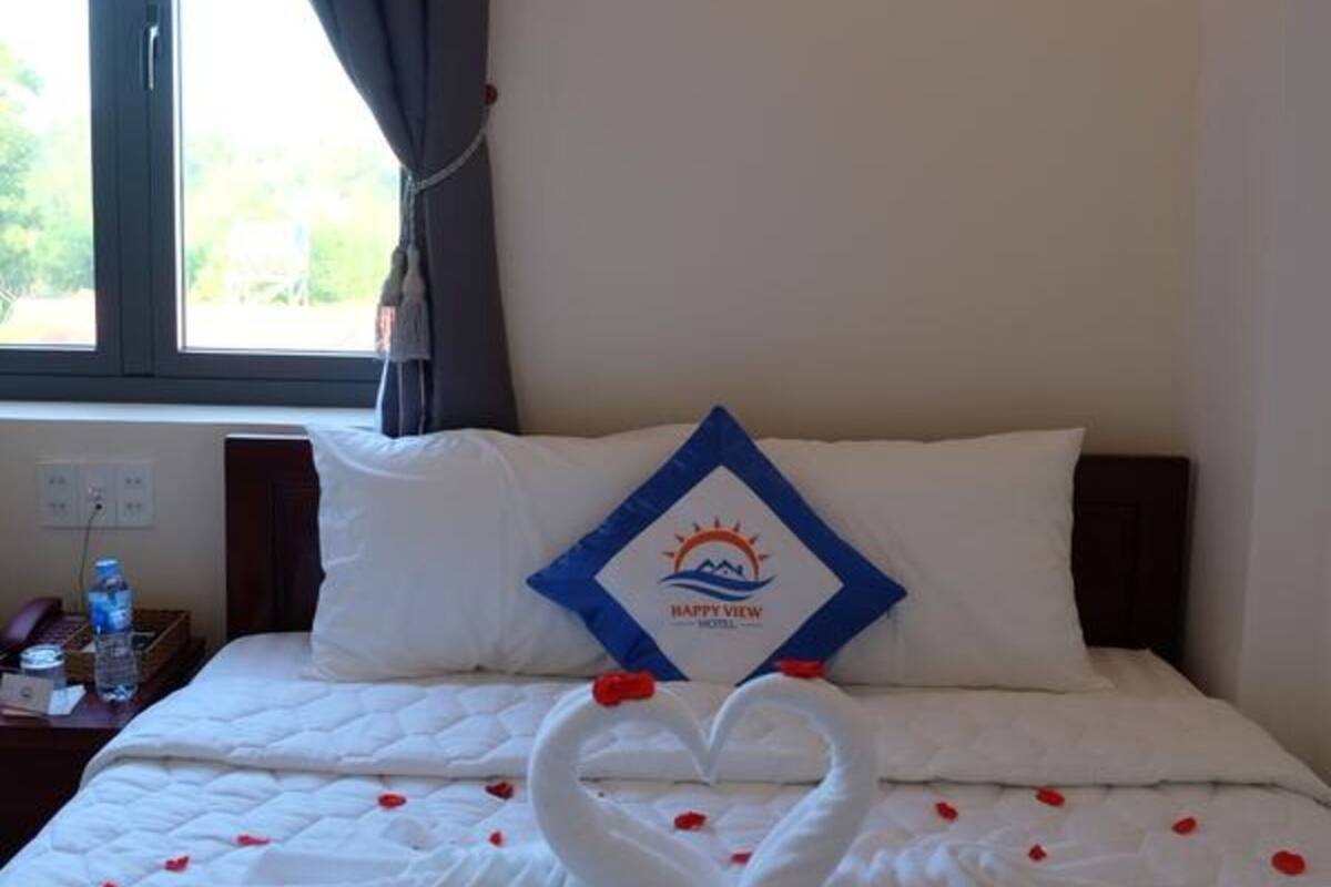A hotel room with a bed, a wall-mounted light, a window with curtains, and a pillow with a logo.