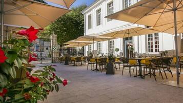 Een terras met tafels en stoelen onder grote parasols, omgeven door een wit gebouw met meerdere ramen.