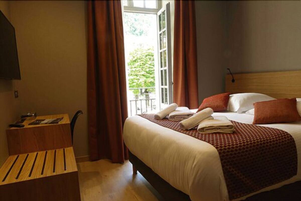 Quarto de hotel com cama, escrivaninha de madeira e janela com cortinas.