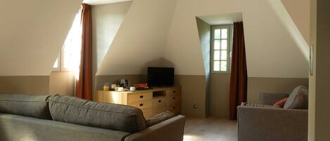 Una habitación moderna y luminosa con un sofá, un sillón más pequeño, un televisor sobre un aparador de madera y una ventana con cortinas.