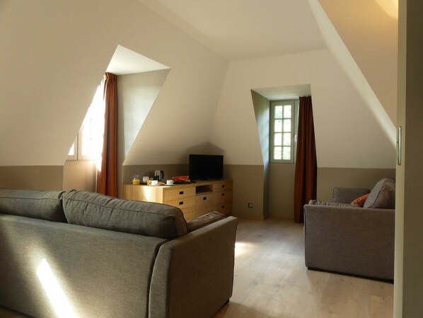 A modern, well-lit room with a sofa, a smaller couch, a TV on a wooden dresser, and a window with curtains. - Le Castel d'Alti (Bagneres-de-Luchon)
