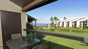 A balcony with a glass table and chairs, overlooking a well-maintained lawn and a row of buildings with a clear view of the ocean. - Hale Kamaole 262  Second floor large condo, Ocean view! (Kihei)